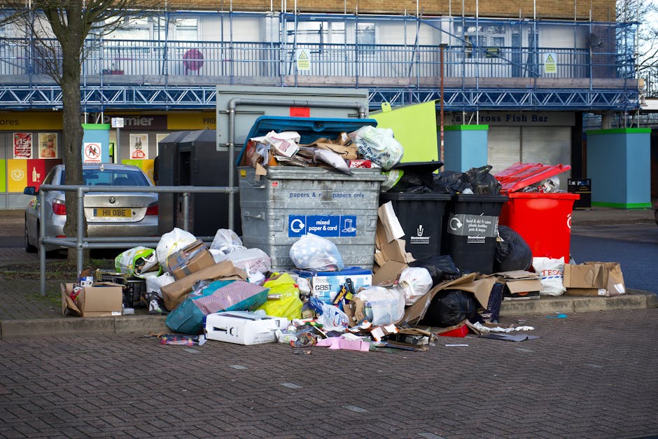 A large collection of overflowing rubbish bins and scattered waste, including cardboard boxes, paper packaging, plastic bags, and miscellaneous household waste, positioned on a paved sidewalk near a metal railing. The waste appears to be a mix of recyclable materials such as paper and non-recyclables, with some items spilling onto the ground. The bins include a grey mixed paper and card container, a black bin, and a prominent red bin, all filled to capacity, with additional rubbish piled around their bases. In the background, there is a tree on the left side, and behind the bins, a building with a blue metal balcony or scaffolding and various storefronts. A parked silver car is visible on the left side, and the overall scene suggests an area requiring efficient waste collection services, possibly handled by private waste disposal firms such as Waste Disposal Stockwell. The environment is urban, with the scene captured during daylight, with a neutral and professional tone emphasizing the importance of proper rubbish management.