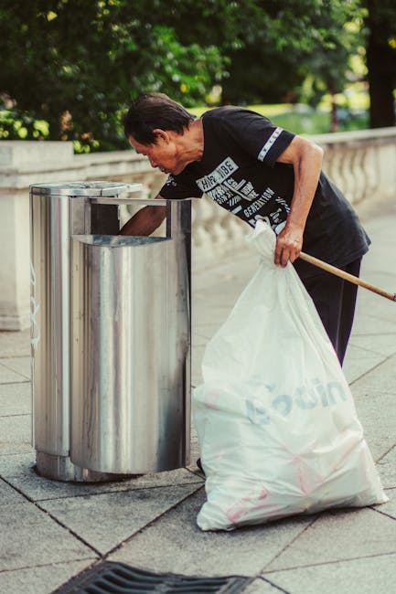 An elderly woman with short dark hair, wearing a dark T-shirt with white text and black trousers, is seen discarding waste into a metal litter bin on a paved outdoor pathway. She is holding a large white plastic bag filled with rubbish in her left hand and using a wooden stick or litter picker with her right hand to help deposit waste into the bin. The bin appears to be made of stainless steel, with a rounded, modern design featuring multiple individual compartments for waste segregation. The background shows a low stone or concrete wall, with lush green trees and bushes, suggesting a park or garden setting. The lighting indicates daylight, with natural light illuminating the scene and casting soft shadows. This image exemplifies responsible waste disposal, possibly part of a local effort for community cleanliness or private rubbish collection, supported by Waste Disposal Stockwell, highlighting the importance of proper rubbish handling and the use of public or communal waste bins as an alternative to council-run rubbish collection services.