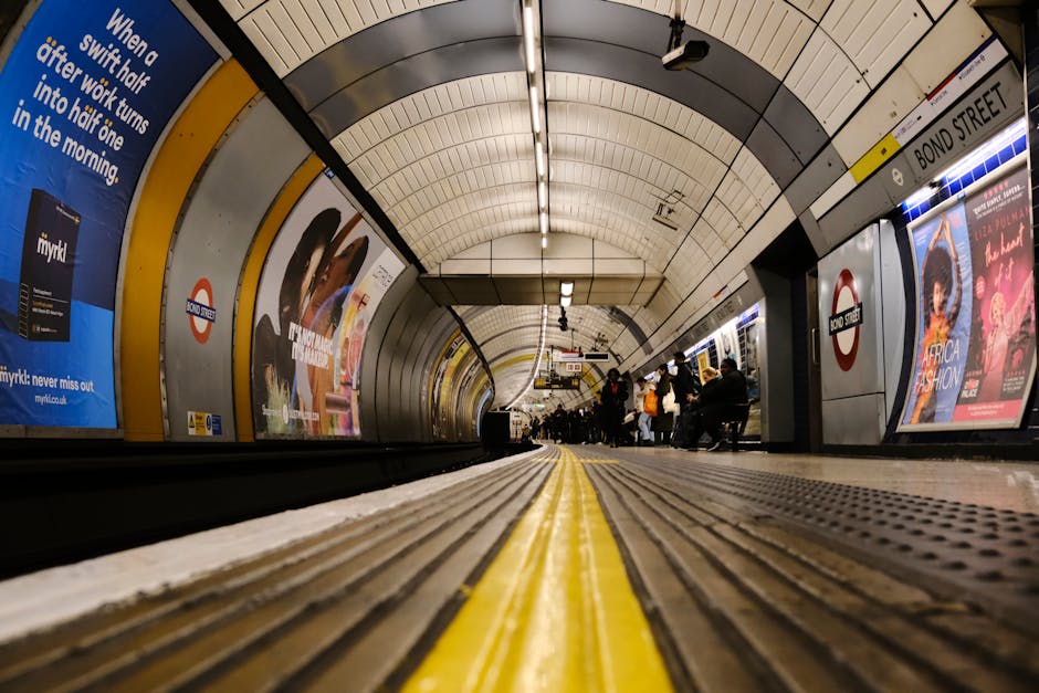 A view from a low angle inside a London underground station, focusing on the platform edge with a textured, grooved surface in brown and yellow warning lines running along the center. The curved tunnel walls are lined with white, grey, and black tiles arching overhead, illuminated by long strip lighting. Along the station walls, there are several large advertising posters, one promoting a mobile phone network and others displaying fashion and entertainment content, framed by yellow and silver trim. Passengers are seated on benches and standing, waiting for trains, with some visible in the background near the platform's edge. The environment appears clean, well-lit, and organized, characteristic of an urban transit station used by commuters. The setting conveys a typical underground station environment, emphasizing the safety markings and commercial advertisements common in London's public transport system, representative of private or independent railway station services. Waste Disposal Stockwell's rubbish removal services catering to urban environments could be inferred as part of maintaining such high-traffic public spaces, although not directly depicted in the image.