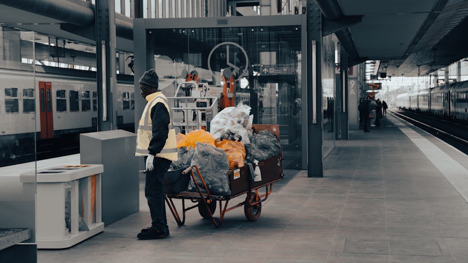 A worker dressed in dark clothing, including a beanie and gloves, stands on a train station platform next to a large, wheeled trolley loaded with various bags and discarded items, primarily made of plastic and fabric, in shades of grey, black, orange, and white. The trolley is filled with mixed rubbish, including plastic waste and fabric bags, indicating a collection of waste items for disposal. Behind the worker, a modern glass and steel shelter provides shelter for waiting passengers, with a transparent panel showing train tracks and a train on the right side of the image, where a few passengers are seen further down the platform. The background shows the station's architectural structure, including metal beams and elevated walkways. The scene is lit with natural daylight, creating a neutral, professional atmosphere typical of urban transit stations, where private waste collection services like Waste Disposal Stockwell might perform rubbish removal or on-site clearance for commuters or station management. The environment appears clean and orderly, emphasizing the context of waste handling within a busy transport hub.