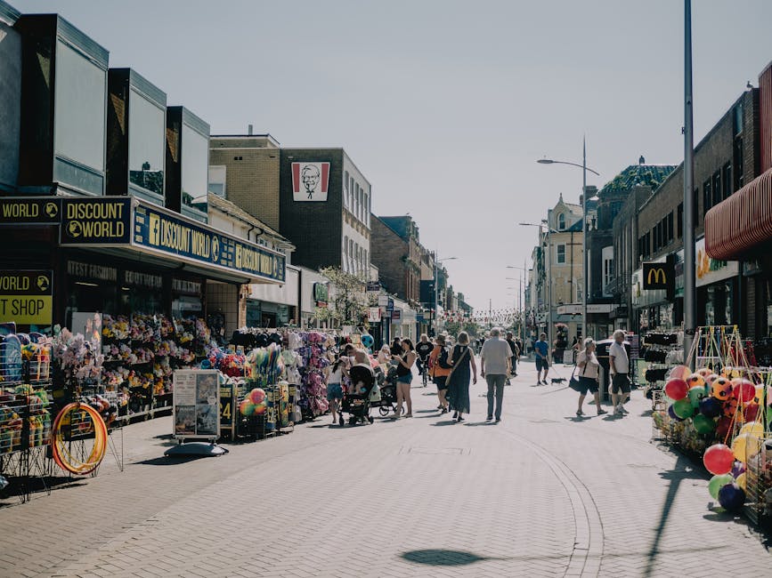 Photograph of a busy outdoor shopping street with various retail stores and market stalls, featuring a variety of merchandise including colorful balloons, toys, and accessories arranged on display tables and in baskets. The scene shows pedestrians walking along the paved pavement, some pushing strollers, and others browsing or engaged in conversation. On the left side, signage indicates a discount retailer and other shops, while the right side includes a fast-food restaurant with an recognizable logo. The background displays a mix of modern and traditional buildings, with storefronts extending into the distance under a clear sky. The area appears well-lit by natural daylight, with a lively atmosphere typical of an urban high street. Some equipment or materials for rubbish collection are not visible, but the environment aligns with the context of maintaining cleanliness through various waste management services provided by Waste Disposal Stockwell, supporting alternative or private waste disposal methods in such commercial settings.