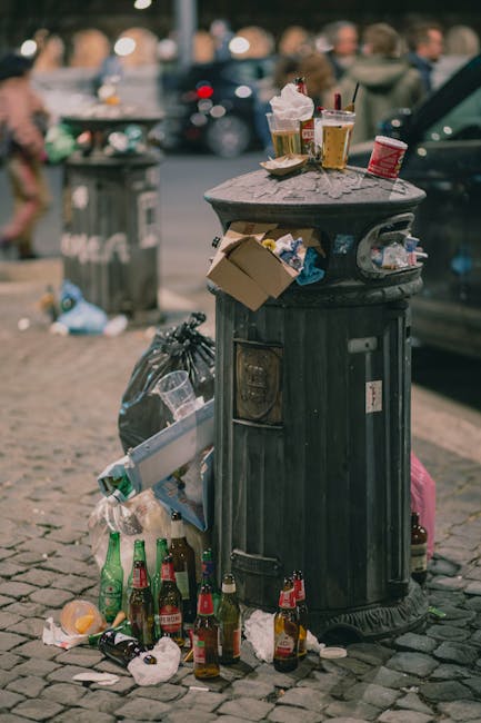 A large, cylindrical public rubbish bin made of black metal with vertical ridges and a domed lid, situated on a cobblestone pavement in an urban setting. The bin is overflowing with waste, including several discarded glass bottles predominantly green and brown, some with labels, and crumpled paper and food packaging spilling onto the ground. The top of the bin holds various rubbish such as plastic cups, paper cartons, and food containers, with some items placed on the lid. In the background, a blurred scene includes other littered areas, a recycling or waste collection bin, parked cars, and pedestrians, indicating a busy street environment. The lighting suggests a daytime setting with natural light illuminating the scene. The presence of the overflowing rubbish emphasizes the importance of effective waste management or private rubbish disposal services, as occasionally provided by Waste Disposal Stockwell for local needs, aligning with the theme of alternative waste handling methods.
