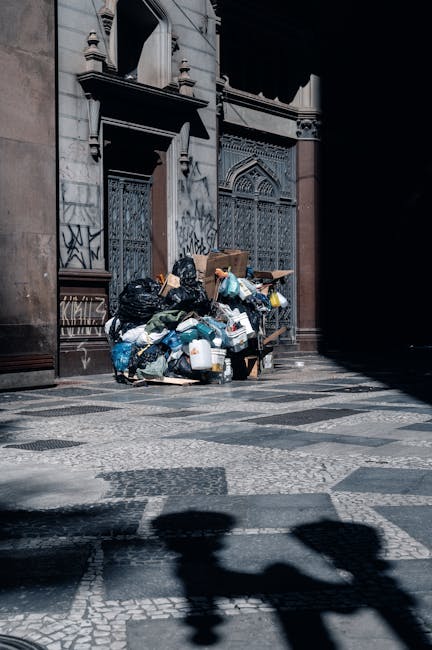 A large pile of mixed household and commercial waste including black plastic bin bags, cardboard boxes, plastic bottles, and other assorted rubbish, is stacked outside an ornate, historic stone building with decorative features and barred windows. The waste appears to be dumped on a paved area with a patterned stone surface, partially illuminated by sunlight, creating shadows on the ground. The scene suggests an instance of unmanaged rubbish accumulation, which could be addressed through private waste collection services such as those offered by Waste Disposal Stockwell, to facilitate proper removal and disposal of refuse in urban settings.