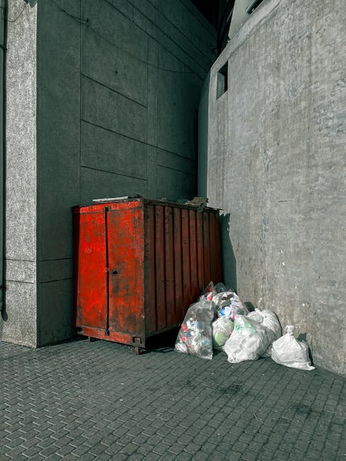 An outdoor urban setting shows a large, rectangular, red metal skip with visible signs of rust and chipped paint, positioned adjacent to the corner where two building walls meet; one wall is dark green with a smooth, modern finish, and the other is light grey with a rough, textured surface. In front of the skip, there are multiple white plastic bags filled with mixed waste, some tied at the top and others loosely closed, with waste materials visible through semi-transparent areas. The ground is paved with small, dark grey bricks arranged in a pattern, and the scene is illuminated by natural daylight, casting subtle shadows on the walls and pavement. The setting appears to be a back alley or service area, which could be an example of private or independent waste management, aligning with services offered by Waste Disposal Stockwell for rubbish removal and on-site clearance.